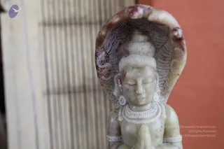 Close-up photograph of a small pale green-white stone carving of a seated deity murti with hands pressed in anjali mudra before the chest. The figure has closed eyes, a tilak mark on the forehead, a high topknot, and a multi-hooded cobra hood rising behind the head. A beaded necklace and earrings are carved on the body. The background shows a blurred wooden latticed panel beside an orange-painted wall.