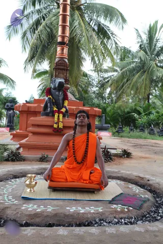 A photograph of a man seated in meditation on a woven mat and orange cushion atop a raised circular stone platform, eyes closed, hands resting on his knees in a meditative posture. He wears an orange-saffron cloth draped over his shoulders and body, a long rudraksha mala necklace, and a red bindi on his forehead, with long dark hair swept back. Behind him stands a black stone Nandi bull statue adorned with a red and yellow flower garland, set on an orange-red pedestal, with a tall copper-clad pillar and coconut palms beyond. A brass kalasha pot sits to the left on the mat, and a small purple-and-green cloth lies to the right. Decorative white floral and swastika-like patterns are drawn on the ground around the platform. Garden statues of standing figures and small shrines appear in the background.
