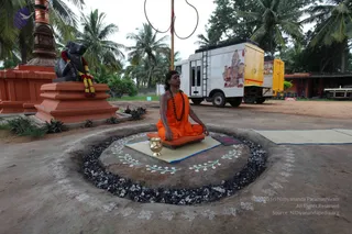 Outdoors in a palm-fringed compound, a man with long dark hair sits cross-legged in meditation on a raised circular earthen platform ringed with dark stones and decorated with white painted floral and swastika motifs. He wears an orange-saffron robe and a long rudraksha mala, with hands resting in his lap. A brass kalasha stands beside him on a low mat. Behind him stands a black Nandi statue garlanded with red and white flowers on a red-painted plinth, a white van with a painted deity mural is parked nearby, and a tall ceremonial pole with a ring stands in the background. The swastika and floral kolam indicate a ritual space; the setting appears to be a ritual or sacred ground.
