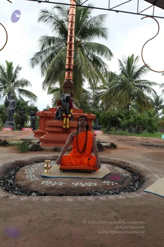 A photograph of an outdoor ritual space with a seated man in orange saffron robes meditating cross-legged on a mat over a circular raised platform ringed with dark gravel. He wears a long dark rudraksha mala and has a red mark on his forehead, eyes closed. A brass kalasha-style pot sits beside him. Behind him stands an orange brick plinth holding a black stone Nandi bull draped with a garland of red, yellow and white flowers, with a tall copper pillar rising above and coconut palms overhead. Several dark stone deity statues stand to the left, and a canopy frame arches overhead. Swirling white kolam-style patterns and mantra-like marks decorate the ground around the platform.