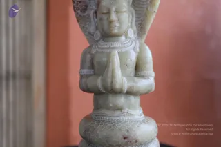 Close-up photograph of a pale green stone carving of a seated deity figure with hands pressed together in anjali mudra, namaskar gesture. The figure wears a beaded necklace, armlets and bangles, with a carved hooded or winged backdrop rising behind the head and a lotus-like base with scale-patterned detailing. A salmon-coloured wall sits in the background. No people are visible; the figure is a sculpted murti, and no a person candidate is present.