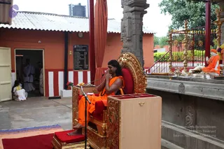 A seated man in saffron-orange robes sits on an ornate gold-and-red throne with a red cushion, hands folded in namaskar at his chest. A microphone stand stands before him and a wooden lectern-like cabinet sits in front. Behind him stands a white-and-gold statue of a seated figure in orange robes with a crown, beside a gold kalasha-style lamp stand and brass vessels with flowers. In the background, a priest in white dhoti and a small seated child in white appear near an orange building with a red-and-white striped band and a plaque. A red-and-gold curtain hangs at left, with a blue-and-orange mat on the ground.