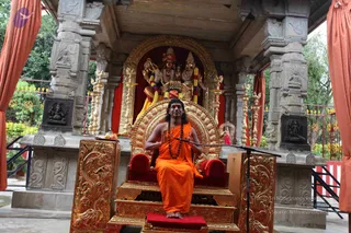 A seated man in saffron-orange robes with long, swept-back dark hair and a rudraksha mala sits barefoot on an ornate golden throne with red cushions, hands raised in a gesture while speaking into a microphone on a stand. Behind him, a small stone-pillared shrine with gold-coloured ornamental lamp stands and a red-curtained backdrop holds a deity murti decorated with a garland of colourful flowers. Orange curtains frame the open-air pavilion, and green trees and a red railing surround it. A fruit offering sits on a tray at the left of the throne.