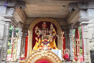 A photograph of an ornate stone shrine with carved pillars and a gold-framed arch. Inside stands a large deity murti of a male and female figure in gold-coloured dress with red and orange cloth, garlanded with multicoloured flower strings. The male figure holds a drum-like object and the female figure holds a flower-garlanded hand. Brass oil lamps on tall stands line both sides. In front, a gold-ornamented throne with a small white-clad figure seated before it, wearing a red and orange turban, appears in the lower foreground. No clearly visible living person is distinct from the murti and the figure at the base, so the count reflects only the seated figure.