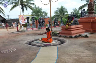 Outdoor photo of a temple-style courtyard with reddish-brown earth. In the centre, a seated figure in orange robes and a rudraksha mala sits cross-legged on a low wooden platform inside a circular stone-edged pit, with one hand extended over a brass kalasha-like vessel and a decorated floor design around it. A long woven mat runs from the pit toward the viewer. Behind, a row of dark stone statues including a Nandi bull with red-and-yellow garland sits on an orange pedestal. Two women in saris, one in a red sari and one in yellow, stand in the background near a roadside sign board with a painted devotional scene. Tall palm trees and a tall decorative lamp post frame the scene.