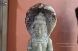 A close-up photograph of a pale green-grey stone carving of a seated deity murti, with eyes closed, hair piled in a high topknot, a snake hood rising behind the head, a multi-strand necklace and hands pressed together in prayer. The background is a warm orange-pink wall, and a blurred dark object sits at the right edge. No people are visible.