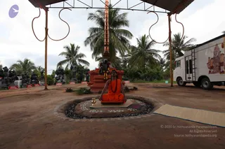 Outdoor photo of a seated figure in orange robes and a red-orange shawl with a rudraksha mala, sitting cross-legged on a cushioned seat within a circular bed of dark gravel and white floral kolam patterns on bare earth. Behind is an orange-red pillared structure with a black bull statue and garlands, a tall copper-coloured pole with a lamp-like top, and a line of dark stone deity murti statues along the left. Coconut palms and a white van with a painted deity mural stand at the right under an overcast sky. A woven mat lies on the ground nearby.