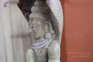 Close-up photograph of a pale grey-green stone carving of a deity, shown in profile with eyes lowered, hair piled in a tall topknot, large ear ornament, and a beaded collar necklace over bare shoulders. Behind the head rises a carved cobra hood with scale-patterned detail. The statue's right hand is raised in a gesture beside the figure, and a red-orange wall lies to the right with a pale blurred background on the left. No living people are visible. The figure is a stone murti, not a person.