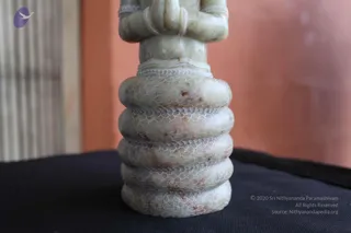 Close-up photograph of a pale grey-white stone sculpture of a seated figure, showing the lower torso with hands joined in front and bangle-like bracelets on the wrists, resting on a stack of five rounded tiers carved with fine scale or lattice patterns. The sculpture stands on a black cloth against a blurred orange wall and a pale vertical grille at left. No people are visible.