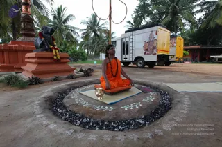 Outdoor photograph of a man seated cross-legged in meditation on a raised circular earthen platform ringed with dark charcoal-like pebbles and decorated with white floral kolam patterns. He wears an orange-saffron robe and a rudraksha mala, with long dark hair swept back, eyes closed and hands resting in his lap. A brass kalasha-style pot sits before him on the mat. Behind him stands a dark Nandi bull statue garlanded with red and orange flowers beside an orange-painted pillar, with a white mobile vehicle and a yellow vehicle parked under coconut palms. A small orange-roofed building is at right. Appearance-based cues of long swept hair, saffron robe and rudraksha mala suggest a person, so the label is given with caution. The sign and layout suggest a meditation or satsang gathering in an ashram setting.