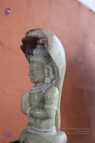 A photograph of a carved greenish stone figure of a seated deity murti with a hooded serpent canopy arching behind its head. The figure has an upswept topknot, ornamented earring and necklace, and bangles, with hands held together in anjali mudra before a coiled serpent base. The background is a flat orange-pink wall.