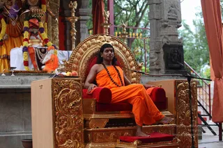 A man in saffron-orange robes sits on an ornate gold-carved throne with red velvet cushions, a rudraksha mala around his neck, and bare feet resting on a red footstool. A microphone stand stands beside him. Behind him are a deity murti draped in marigold and multicolored flower garlands, a seated orange-robed figure, brass lamps and a kalasha-style vessel, with a stone pillar and green trees in the background. The setting appears to be an outdoor open pavilion used for satsang. Appearance-based label: a person, seated on the throne, identified by long swept hair, saffron clothing and rudraksha mala.