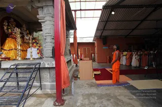A photograph of an open-sided temple courtyard with a corrugated roof and red steel posts. On the left, a shrine holds deity murti in gold and orange garments with flower garlands, flanked by tall brass oil lamps. A man in a saffron-orange robe stands barefoot on a red carpet, facing the shrine with hands raised in devotion. Behind him, a row of about a dozen people in white and orange clothes stands in line, some with hands folded. A wooden lectern, a low red mat and a patterned rug lie on the floor, and a wrought-iron shelf stands at the left edge.