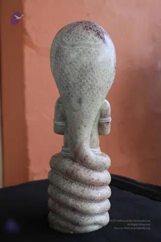A pale green-grey stone sculpture of a seated figure viewed from behind, with a multi-hooded serpent canopy carved in fine scale patterns rising above its head. The figure's shoulders and arms are visible with bead-patterned armbands, and the base is a coiled serpent body with red-brown veining in the stone. The sculpture rests on a black cloth against a terracotta-coloured wall. No physical people are visible, and no a person candidate is present; the figure is a stone statue.