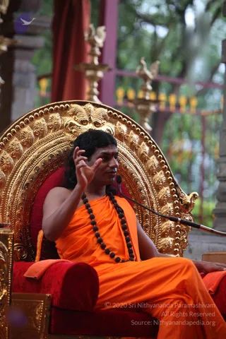 A photograph of a man with long dark hair seated on an ornate gold throne with a red cushioned seat and red velvet backrest. He wears a saffron-orange draped garment and a long necklace of dark rudraksha beads, with a small red mark on his forehead. He raises one hand in a gesture while speaking into a microphone on a stand. Behind him are brass oil lamps with bird-shaped finials, red drapes and green trees. A translucent purple bird logo appears in the upper left. The person is an appearance-based a person candidate given the long swept hair, saffron clothing, rudraksha mala and throne, presented as a teacher giving a satsang talk.