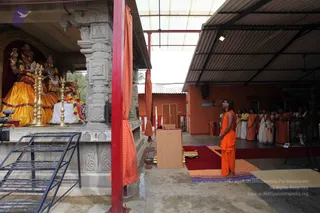 A photograph taken inside a covered open-sided temple courtyard with an orange-painted wall and corrugated roof. On the left, a shrine holds seated deity murti in gold and orange garments with floral garlands, flanked by tall brass oil lamps. In the centre-right, a man in an orange dhoti and orange shawl with a garland stands on a red carpet beside a wooden lectern. Behind him, a line of about a dozen people in white and orange garments stands along the wall, some with hands folded. Two white-clad figures in the foreground-right have their hands together. A red pillar and orange cloth hangings frame the scene.