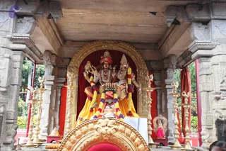 A photograph of an ornate stone shrine with carved pillars and a gold-framed arch. Inside, two large seated deity murtis are garlanded with yellow, orange and purple flower garlands; the central figure wears a red and orange crown and a multi-strand bead necklace, while the figure to the right wears a gold crown and gold necklace. Both are draped in yellow and orange cloth. Brass oil lamps stand on either side, and a golden ornate throne-like seat with a red cushion sits in the foreground. A small pink-clad figurine is placed to the right. No living people are clearly visible; a partial figure appears at the lower right edge.