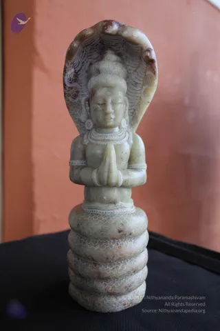 A carved pale green-grey stone statue of a seated deity in anjali mudra, hands pressed together at the chest, with eyes closed and a topknot hairstyle. The figure wears a layered necklace, earrings and bangles, and a cobra hood with scale-patterned carving rises behind the head. The statue rests on a coiled serpent base with scale-patterned coils, set on a black cloth against an orange wall. This is a statue, not a person, and no candidate is present in the image.