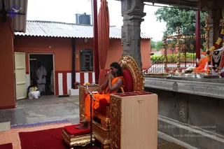 A photograph of an outdoor temple-courtyard setting. A man with long dark hair, wearing an orange-saffron robe, sits on an ornate gold-and-red throne with his palms pressed together in a namaste gesture, bare feet resting on a red cushioned footstool. A microphone stands before him and a wooden lectern with red top sits in front. To the right, a deity murti adorned with a flower garland and a beaded necklace is seated on a raised platform beside brass lamps and a kalasha, with another orange-robed figure seated nearby with hands folded. In the background, a man in white dhoti and a person in white clothes stand in an arched doorway of an orange building with a striped red-and-white wall and a corrugated roof, and a seated figure in white sits near the entrance. The tall temple pillars and brass lamps frame the scene. Whether the seated speaker is a person is uncertain from appearance alone, so he is described by role.