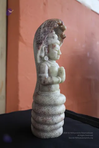 A carved pale stone statue of a seated deity murti with palms pressed together in anjali mudra, placed on a coiled serpent base with a hooded cobra canopy behind the head. The stone has a greenish-grey tone with reddish veining, and fine incised scale patterns cover the cobra and coils. Hair is piled in a high topknot, with a round earring and a beaded necklace visible. The statue sits on a dark cloth against an orange-painted wall.