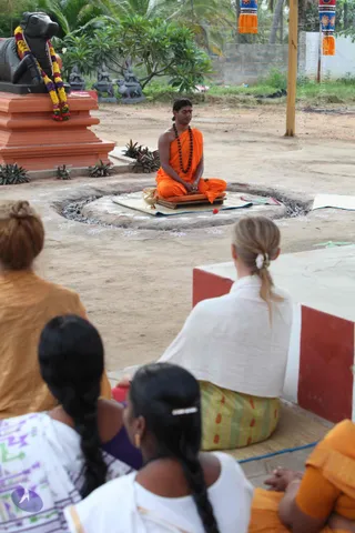 Outdoor photo of a seated man in orange robes and a rudraksha mala meditating with eyes closed on a low wooden seat and mat beside a circular fire pit with ash and a brass vessel. Behind him stands a black Nandi bull statue with a garland of marigolds and purple flowers on an orange pedestal, with small deity statues and frangipani plants in the background. Hanging orange and colourful cloth banners frame the upper right. Several seated people face him from the foreground, including a woman in white with blonde hair tied back, a woman in orange, and two women with long braids, one wearing white.