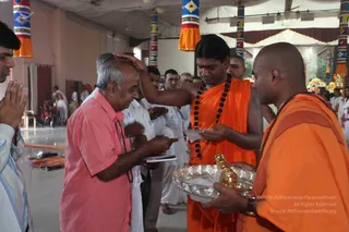 A photograph inside a hall with white ceiling fabric and colourful striped banners hanging from above. A man in a pink short-sleeved shirt with grey hair stands with palms pressed together while a younger priest in an orange robe and a long rudraksha mala places his hand on the man's head, apparently giving a blessing. The priest holds a paper, and beside him a bald attendant in orange with a rudraksha mala holds a silver tray containing a small gold-coloured ritual object. Behind them, men in white dhotis and orange-clad figures stand in a line, and flower decorations appear at the far right.