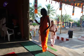 A photograph of an outdoor ashram setting under a covered walkway hung with colourful cylindrical banners, flags and a garland-like ornament. A figure with long dark swept-back hair, a rudraksha mala and a saffron-orange garment walks barefoot across a green mat at a doorway, one hand raised. Beside and slightly behind, a shaved-headed man in white dhoti and shawl with red border walks along the steps. Stone sculptures line the courtyard in the background, and a palm-lined ground and white building are visible. Swamiji (probable) appears in the saffron robe with long hair and rudraksha mala, walking as part of a procession or pada puja event.
