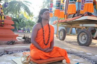 A photograph of a man seated cross-legged on a cushion placed on a mat outdoors, eyes closed, hands folded in his lap in meditation. He wears a saffron-orange robe draped over his shoulders and a long strand of dark rudraksha beads around his neck, with a red bindi on his forehead and long hair swept back. Beside him sits a polished brass kalasha-style vessel with a spout. Behind him stands a wooden cart with orange and green fabric hangings and garlands, under palm trees, with a small orange shrine structure at left. Several bright lamps and flower garlands are visible in the setting. The man is seated and centered, the central figure of a ritual scene.