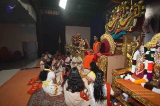 A seated man in an orange-saffron robe sits on a red-cushioned golden throne on a raised dais, with a rudraksha-style bead necklace visible, while a group of about a dozen women and girls in white and cream saris and dhotis sit on orange mats before him holding brass plates and offerings. Behind the throne, a large floral garland of marigolds and green leaves drapes over deity murtis in an ornate shrine. A gold-framed ratha-style seat stands at right beside a decorated deity figure with garlands. The setting is a stage-like hall with a dark curtained backdrop, orange floor covering and a patterned carpet. The scene appears to be a padapuja gathering in progress.