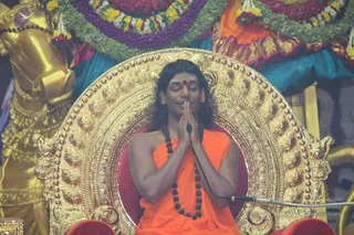 A photograph of a man seated on an ornate gold throne with red cushions, eyes closed and palms pressed together in namaskar. He wears an orange-saffron cloth over his shoulders, a dark rudraksha mala draped across his chest, and a red bindi on his forehead. A microphone stands before him. Behind the throne are a gilded horse-head sculpture and garlands of purple, red and yellow flowers over teal and orange drapery. a person (appearance-based: rudraksha mala, saffron clothing, ornate throne, seated in devotional posture) seated as the speaker.