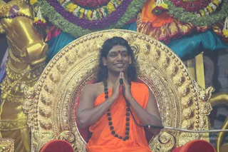 A man with long dark hair and a red bindi sits in an ornate gold throne with carved scrollwork and studded rims, palms pressed together in greeting, smiling toward the audience. He wears an orange saffron robe and a long dark rudraksha mala, with a microphone stand beside him. Behind him are a golden animal figure at left, garlands of yellow, orange and purple flowers, and teal and red drapery. Swamiji (probable): a long-haired figure in saffron with rudraksha beads seated on a throne at a satsang, identified by the page caption as the speaker.