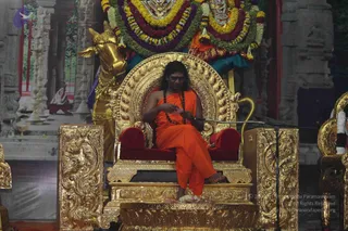 A seated figure on an ornate golden throne with red velvet cushions, dressed in an orange saffron garment with a rudraksha mala draped across the chest, holding a small pink paper or card in both hands while speaking into a microphone on a stand. Behind the throne stands a golden bull statue, and above are large garlands of yellow, orange, purple and green flowers framing deity murti images. Flower petals lie on the golden platform in front.