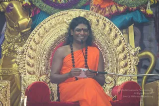 A smiling man with long dark swept-back hair sits on an ornate gold throne with red velvet cushions, wearing an orange saffron wrap and a rudraksha mala with dark beads. He has a red bindi on his forehead and holds a small white card in his hands, with a microphone on a stand in front of him. Behind the throne are draped teal and purple fabrics decorated with yellow and purple flower garlands, and a golden figure stands at the left edge. Swamiji (probable) is seated centrally as the speaker during a satsang discourse, identified by the orange wrap, long hair, and rudraksha mala.