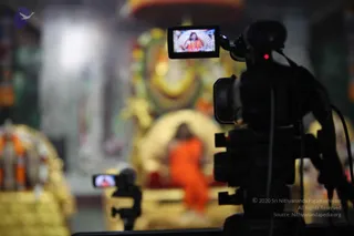 A video camera in the right foreground, with a small monitor on its side showing a live image of a man in orange robes seated before a backdrop. Behind it, a blurred temple interior shows a seated figure in orange robes on a gold and red throne, framed by gold garlands, flowers and a golden canopy. A second, smaller camera stands at left, its screen showing the same scene. The people on the screens and the blurred figure are depictions or distant presenters; no individual physical person is clearly in the foreground. The scene shows a satsang discourse being filmed.