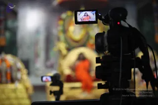 A video camera on a tripod in the foreground records a satsang hall. Its small monitor shows a figure in orange robes seated on a raised golden throne. In the blurred background, a seated figure in an orange robe sits before a golden arch and ornate golden decorations, with garlands and flowers around the altar area. A second small camera stands in the lower left. The scene suggests a live filmed talk, though the blurred figures are too indistinct to count as clearly visible people beyond the orange-robed seated figure.