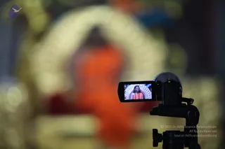 A video camera on a black tripod in the right foreground records a seated figure in the blurred background. The camera's flip-out screen shows a close view of a seated person in orange robes wearing a rudraksha mala, in front of an ornate white and gold throne with a gold arch. The blurred background is dominated by orange and gold tones, with a red cloth near the bottom. The camera points toward a seated figure in saffron-orange robes before a golden backdrop.