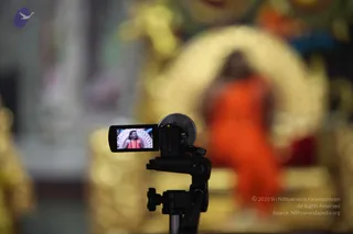 A video camera on a black tripod is in the foreground, its flip-out screen showing a close view of a figure in orange robes with a rudraksha-style necklace in front of a gold-framed backdrop. Behind the camera, blurred, a person in an orange-saffron robe sits or stands before a large gold arched throne-like structure with ornate gold decor. The background is out of focus with gold and yellow ornamentation. a person: the figure on the camera screen and the blurred orange-robed figure have a compatible presentation, with saffron-orange robes and a rudraksha-style necklace, seated before the golden throne.