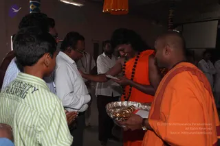 Photograph of a pada-puja moment inside a hall with a screen and banners at the top. A long-haired man in an orange robe and black rudraksha mala stands centre, smiling and reaching toward a man in a white shirt who holds a folded paper. On the right, a shaved-headed man in an orange robe holds a silver tray with brass items. Around them, several men in white shirts and dhotis and a boy in a green striped shirt watch, some with hands joined. a person: the long-haired figure in orange with rudraksha mala is a candidate, based on hair, orange clothing and mala cues, shown as the one receiving the devotee's offering.