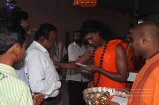 A photograph of a padapuja-style encounter in an ashram hall with pink walls and a hanging orange lamp. A figure with long, swept-back dark hair, orange saffron garments and a dark rudraksha mala stands at centre, leaning forward and handing a folded white paper to a man in a white shirt and spectacles who bows with hands clasped near his chest. Behind them a bearded man in white shirt and dark trousers and other onlookers stand, and a silver tray holds gold ornaments at lower right. A younger person in a green striped shirt and a bald man in orange robes with a rudraksha mala are in the foreground. a person is the central figure in orange garments with a rudraksha mala and flowing hair.