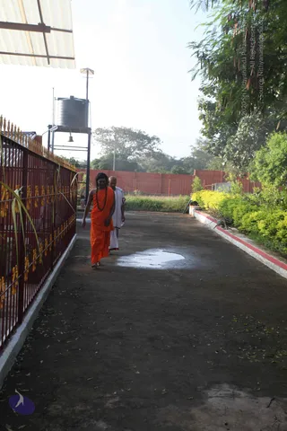Outdoor ashram path on a sunny morning with a wet puddle on the dark pavement. A long-haired man in an orange saffron wrap and a rudraksha mala walks barefoot toward the camera, followed by a shaved-headed man in white dhoti and shirt. Red iron railing with gold-tipped spikes lines the left, a silver water tank on a stand and a bell hang behind, a red boundary wall, green shrubs and trees stand to the right.