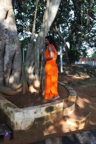 A photograph of a man in a flowing saffron-orange robe standing barefoot beside the large trunk of a banyan tree with dangling aerial roots. He stands on the raised stone platform around the tree base, facing left in a quiet, still pose, with long dark hair falling past his shoulders. Sunlight and shadow play across the bare earth and stone steps, and the sprawling canopy fills the background.