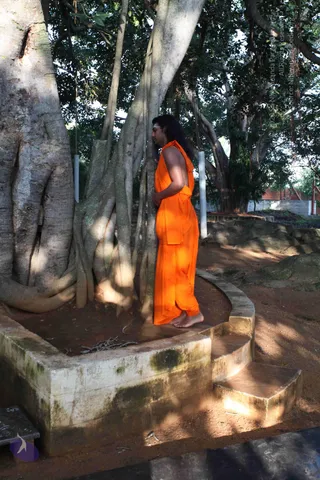 A photograph of a single man standing barefoot in profile beside a large banyan tree with many aerial roots, on a circular raised stone platform with steps. He wears a loose orange saffron garment draped over one shoulder and wrapped around the lower body, and has long dark hair swept back and a tilak on his forehead. He stands with his hands clasped at his waist, facing the trunk in quiet devotion. Loose sticks lie on the earth in the platform, dappled sunlight falls across the ground, and a white wall and rocks are visible behind.