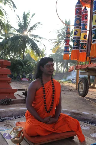 Outdoor photograph of a long-haired man in saffron-orange robes seated cross-legged on a cushion with eyes closed in meditation, hands resting in his lap, a red tilak mark on his forehead and a dark rudraksha mala around his neck. Behind him are palm trees, a decorated wooden chariot with colourful striped and orange-fringed hangings, and a red stone shrine structure to the left. A brass pot stands in the lower left foreground on a woven mat with white floral kolam patterns on the ground.