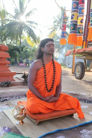 A man with long, swept-back dark hair sits cross-legged with eyes closed on a low wooden seat with an orange cushion, on a sandy ground outdoors under palm trees in bright sunlight. He wears a saffron-orange shawl draped over his body and a long black rudraksha mala around his neck, with a red bindi on his forehead, and his hands rest in his lap. Beside him on a woven mat stands a polished brass kalasha-style vessel with a spout. Behind him a wooden cart with orange-and-blue decorated tassels and a painted fringe is visible, with a faint wisp of smoke. Swamiji (probable) is identified by the long swept-back hair, saffron shawl and rudraksha mala, and the page places him at the Pada Puja event.