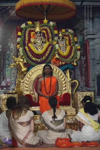 A photograph of a Pada Puja ceremony in a temple-style hall. A man with long dark hair, a red tilak and a black rudraksha mala sits eyes-closed on an ornate gold throne with red cushions, wearing a saffron-orange shawl over bare shoulders, hands folded in his lap. Four devotees in white and cream clothes with gold borders sit on the floor with their backs to the camera, facing him. Behind the throne are two deity murti adorned with bright garlands of yellow, red and green flowers, a golden bull figure at left, and a gold parasol overhead. Saffron cloth sits folded on the floor at lower right. A painted temple backdrop covers the wall.