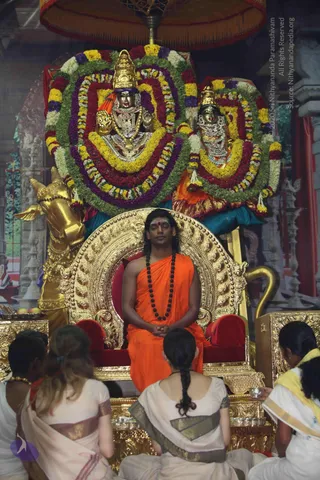 A seated man in an orange saffron robe and a long black rudraksha mala sits with eyes closed and hands folded on a gilded throne with red cushions, a red tilak on his forehead. Before him, four women in white and cream saris sit with their backs to the camera, one in a yellow-bordered sari at right. Behind the throne, two golden deity murtis with crowns are draped in dense garlands of yellow, red, purple and green flowers, with a golden bull figure at left and a golden canopy above. Small brass vessels sit on the gilded platform. The scene suggests a pada puja offering to the seated figure.