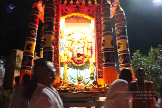 Night procession photograph of a decorated ratha or palanquin-style shrine with red, gold and black-patterned pillars and orange fringe. Inside the shrine stands a deity murti in a blue-white cloth, draped in yellow and orange marigold garlands and a red-pink garland. Brass offering trays with fruit and flowers sit on the cart in front of it. In the foreground, bald men in white and saffron-orange robes walk beside the cart; one wears a rudraksha mala and has a shaved head, and another in orange garments turns toward the shrine. Lush bushes and dark night sky frame the scene.