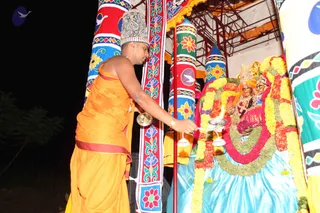 A photograph taken at night of a priest in an orange-saffron dhoti and shawl, wearing a decorated silver-and-gold crown, lighting an oil lamp with a flame before a deity murti. The murti is adorned with heavy yellow and white marigold garlands, red flower garlands and a green leaf garland, with a blue satin cloth beneath. Behind is a colourful painted pillar with floral motifs and a bird design, and a metal scaffold frame with orange fabric overhead. The priest appears to be performing aarti or deepa puja; one person is visible.