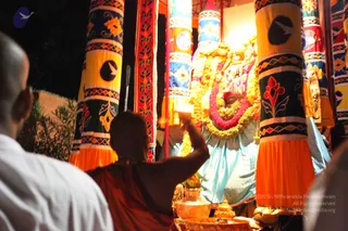 A night-time photograph of a deity murti decorated with marigold and red-pink flower garlands, set within tall banner-pillars with yellow, orange, blue and dark-green fabric panels and embroidered floral motifs. In the foreground, a shaved-headed figure in an orange-saffron robe stands with raised right arm, facing the decorated murti, with a brass vessel and small offerings in front. A man in a white shirt stands at the left edge, partly visible. Oil lamps glow behind the decorations.