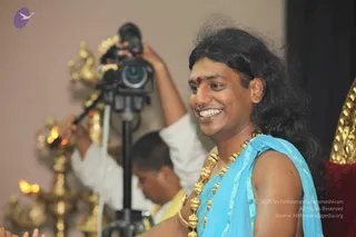 A smiling man with long, swept-back black hair and a red bindi sits at the right of the frame, wearing a light blue shawl over his bare shoulder and a heavy ceremonial gold necklace of large beads. A cameraman in white with a video camera on a tripod stands behind him, and brass oil lamps with lit flames sit in the left background. The man has long hair, a gold necklace and a shawl, consistent with the a person cues, but the appearance is not conclusive, so this is marked unsure. Golden ornamental elements and a red cushion appear at the right edge.
